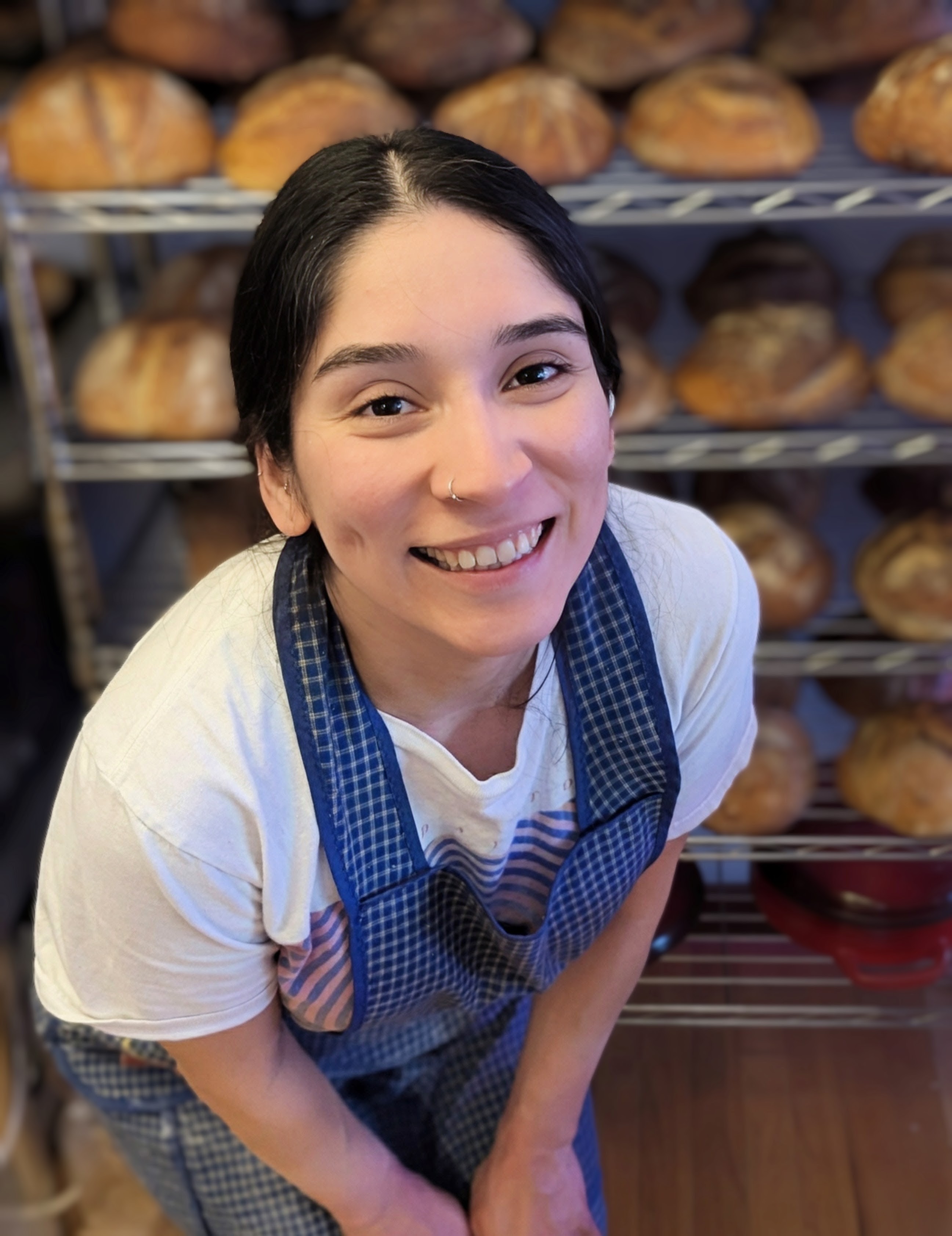 Sarahi in the bakery, racks of fresh loaves behind her